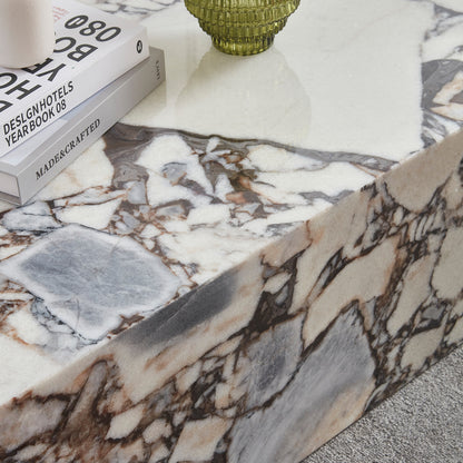 Solene 100cm Coffee Table in Blue Galaxy, a polished marble piece with cream, gray, and rust veining. Topped with design books and a green textured glass vessel, on a light carpeted floor.