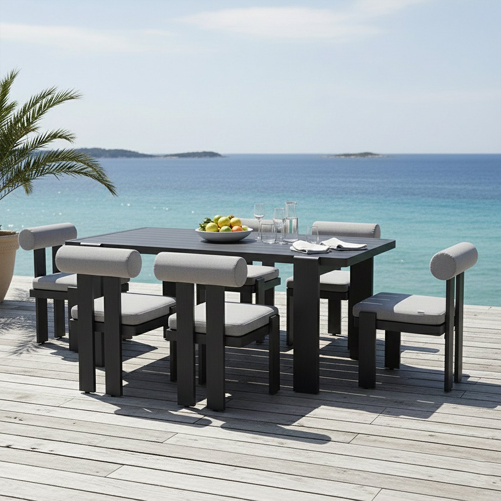 Positano 6 Seater Outdoor Dining Set in Black & Grey on a deck, featuring a rectangular table, six padded chairs, a fruit bowl, glassware, and a potted palm, overlooking the sea.