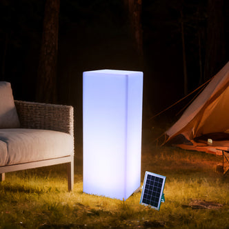 80cm Outdoor Solar LED Column Light glowing bluish-white, stands near a solar panel, beside a woven-frame lounge chair on grass, with a tent and trees in the nighttime background.