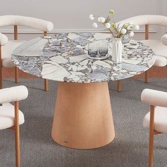 Dina 120cm Round Marble Dining Table in Blue Galaxy Natural with a white-gray marble top and light-wood base, surrounded by boucle-upholstered chairs, a vase with flowers, and glasses on the table.