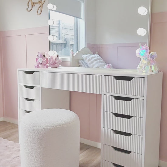 
A cinematic video of a white vanity desk with ridged drawers, featuring a large mirror with light bulbs. The video opens with a close-up of a hand pulling out a drawer to reveal a set of colorful pens. The camera then pans out to show the full vanity setup, which includes a fluffy white stool with a gold base. The vanity is placed in a child's bedroom with light pink walls, a pink fuzzy rug, and stuffed animal decorations, creating a fun and youthful atmosphere.