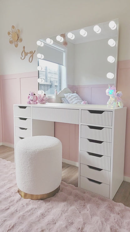 
A cinematic video of a white vanity desk with ridged drawers, featuring a large mirror with light bulbs. The video opens with a close-up of a hand pulling out a drawer to reveal a set of colorful pens. The camera then pans out to show the full vanity setup, which includes a fluffy white stool with a gold base. The vanity is placed in a child's bedroom with light pink walls, a pink fuzzy rug, and stuffed animal decorations, creating a fun and youthful atmosphere.