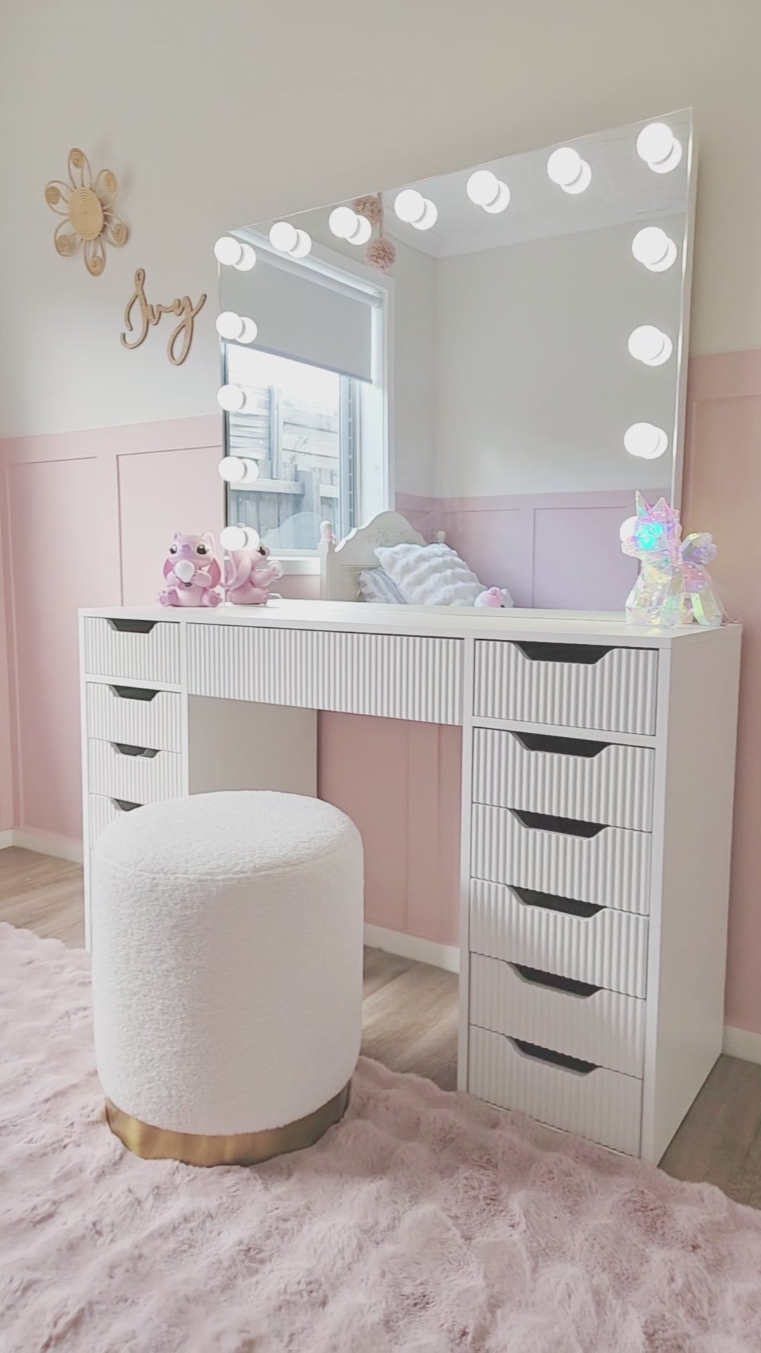 
A cinematic video of a white vanity desk with ridged drawers, featuring a large mirror with light bulbs. The video opens with a close-up of a hand pulling out a drawer to reveal a set of colorful pens. The camera then pans out to show the full vanity setup, which includes a fluffy white stool with a gold base. The vanity is placed in a child's bedroom with light pink walls, a pink fuzzy rug, and stuffed animal decorations, creating a fun and youthful atmosphere.