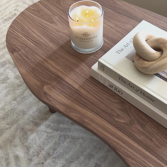 
A video showing a beautifully styled living room. The camera moves from a close-up of a lit candle on a walnut wood coffee table to reveal two decorative books, a carved wooden chain link, and a mushroom-shaped brass lamp. The unique coffee table features a freeform, organic shape. The shot then pans up to show a gray sofa with a textured blanket, highlighting a cozy and neutral aesthetic.