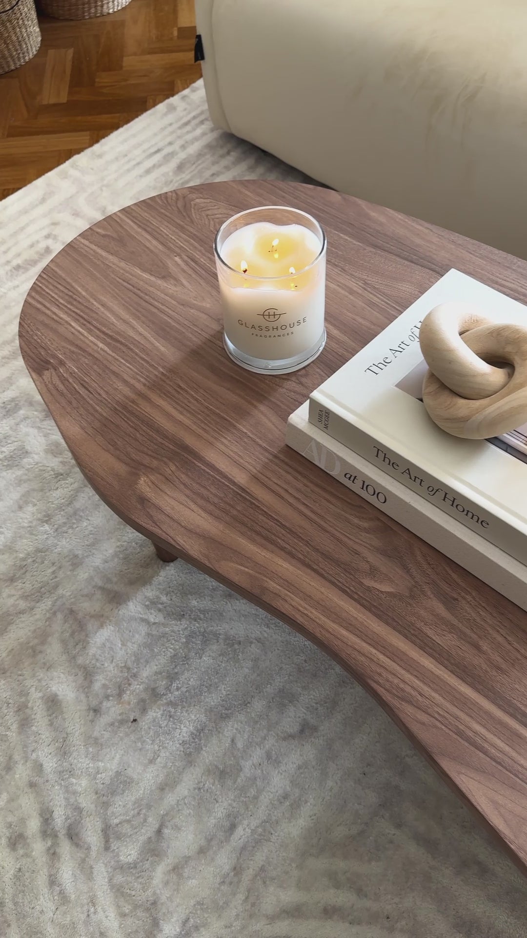 
A video showing a beautifully styled living room. The camera moves from a close-up of a lit candle on a walnut wood coffee table to reveal two decorative books, a carved wooden chain link, and a mushroom-shaped brass lamp. The unique coffee table features a freeform, organic shape. The shot then pans up to show a gray sofa with a textured blanket, highlighting a cozy and neutral aesthetic.