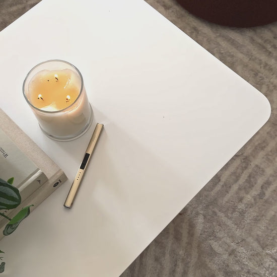 
A close-up of a modern, white square coffee table with softly rounded edges. The camera pans over the tabletop, showing a decorative stack of books with a small houseplant on top, a lit candle, and a gold table lighter. A small brass lamp with a burlap shade sits on the books, adding a warm glow to the scene. The shot then widens to show the table next to a neutral-toned sofa and a fluffy, brown ottoman, highlighting a cozy, contemporary living room setup.