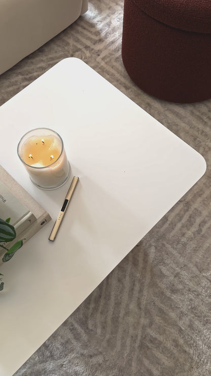 
A close-up of a modern, white square coffee table with softly rounded edges. The camera pans over the tabletop, showing a decorative stack of books with a small houseplant on top, a lit candle, and a gold table lighter. A small brass lamp with a burlap shade sits on the books, adding a warm glow to the scene. The shot then widens to show the table next to a neutral-toned sofa and a fluffy, brown ottoman, highlighting a cozy, contemporary living room setup.