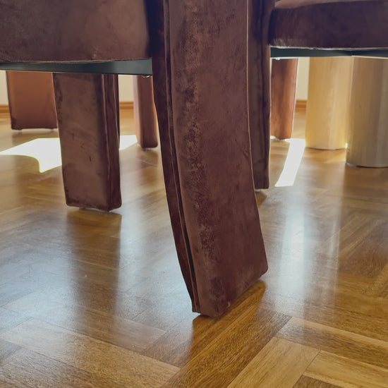 
A slow, cinematic close-up of a modern living room armchair with a unique open-back design. The chair has rounded, curved arms and is upholstered in a plush, rust-colored, velvety fabric. The camera moves down to the chair's base, showing its unique, curved feet. The shot highlights the chair's soft texture and elegant, contemporary form.