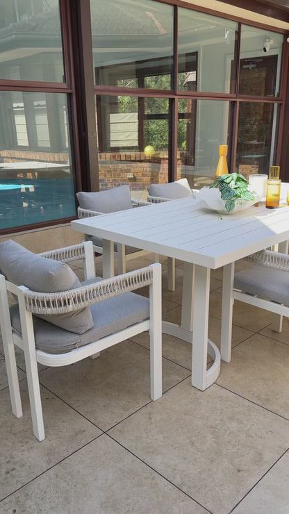 
A wide shot of a modern outdoor dining set. The set includes a large, white rectangular table with a unique half-circle base and multiple chairs. The chairs have white frames, thick gray cushions, and intricate white rope detailing on the backrests. The table is styled with a white bowl of greenery, a yellow vase, and two glasses with a golden-hued drink. The furniture is set on a tiled patio, with a swimming pool visible in the background.
