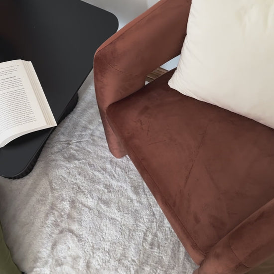 
A video of a modern living room. The camera starts with a close-up of an open book on a black rectangular coffee table with softly rounded edges. The camera then pans out to reveal the full table, which has a unique base with ridged, half-circle-shaped legs. The table is styled with a book stack, a black candle holder, and a white, textured vase with yellow tulips. The surrounding area features a brown velvet armchair and a fluffy white rug with green and brown pillows, creating a warm, cozy aesthetic.