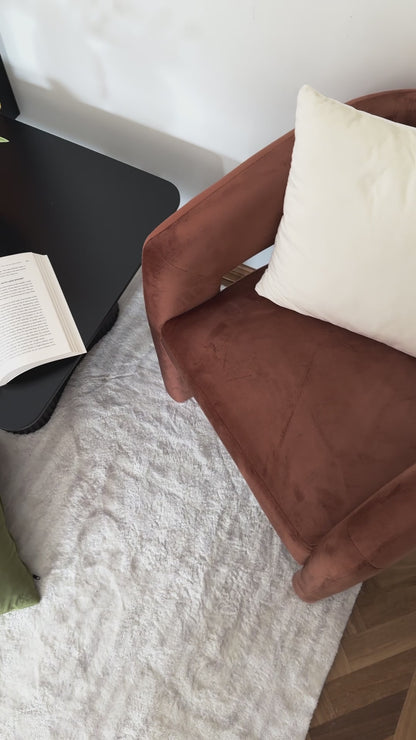 
A video of a modern living room. The camera starts with a close-up of an open book on a black rectangular coffee table with softly rounded edges. The camera then pans out to reveal the full table, which has a unique base with ridged, half-circle-shaped legs. The table is styled with a book stack, a black candle holder, and a white, textured vase with yellow tulips. The surrounding area features a brown velvet armchair and a fluffy white rug with green and brown pillows, creating a warm, cozy aesthetic.