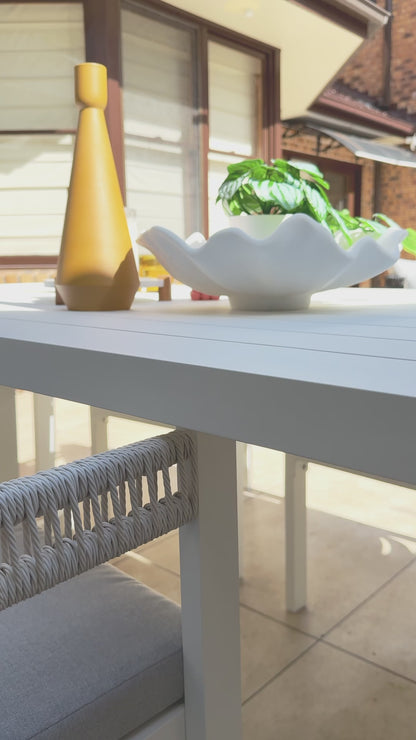 
A close-up of a white, modern outdoor dining table decorated with a gold vase and a wavy white bowl. The camera pans slowly to a chair with a white frame and thick, woven ropes forming the backrest. The seat and back cushions are a comfortable gray, highlighting the elegant design of the outdoor furniture set. The background shows a patio with large windows and a covered seating area.