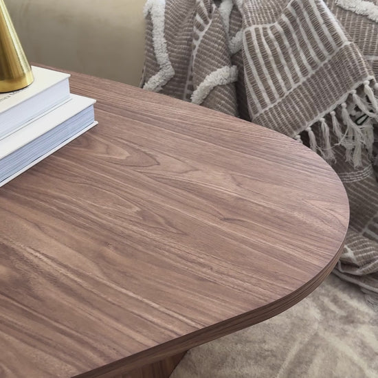 
A close-up video of a walnut wood coffee table with a unique, rounded, freeform shape. The tabletop's grain is visible as the camera pans slowly across its surface. The table is styled with a lit candle, a stack of books, a carved wooden link, and a mushroom-shaped brass lamp. The shot then widens to reveal a cream-colored sofa with throw pillows and a textured blanket, completing the cozy, modern living room scene.