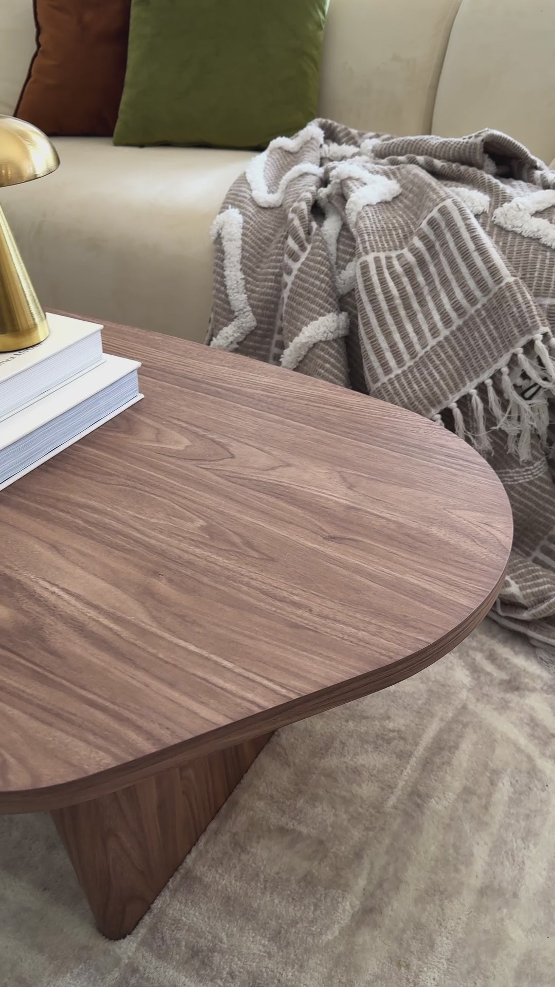 
A close-up video of a walnut wood coffee table with a unique, rounded, freeform shape. The tabletop's grain is visible as the camera pans slowly across its surface. The table is styled with a lit candle, a stack of books, a carved wooden link, and a mushroom-shaped brass lamp. The shot then widens to reveal a cream-colored sofa with throw pillows and a textured blanket, completing the cozy, modern living room scene.
