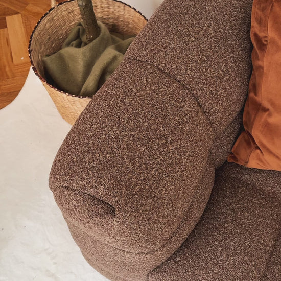 
A cinematic close-up of a brown, textured modular sofa, highlighting its tufted design and soft, boucle fabric. The camera focuses on two decorative pillows resting against the sofa's armrest: a solid burnt orange pillow and a light-colored pillow with an abstract, squiggly brown pattern. The shot captures the inviting texture and cozy aesthetic of the furniture.
