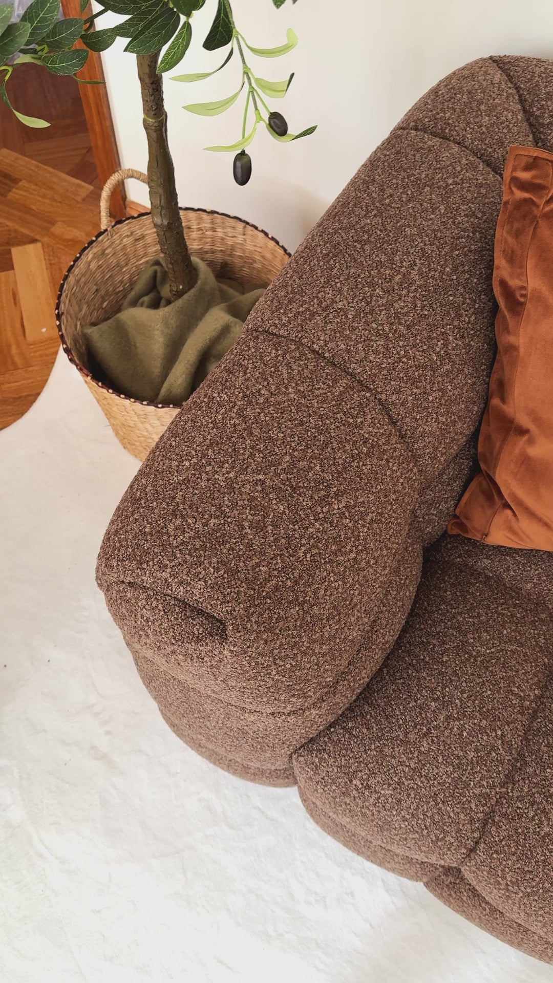 
A cinematic close-up of a brown, textured modular sofa, highlighting its tufted design and soft, boucle fabric. The camera focuses on two decorative pillows resting against the sofa's armrest: a solid burnt orange pillow and a light-colored pillow with an abstract, squiggly brown pattern. The shot captures the inviting texture and cozy aesthetic of the furniture.
