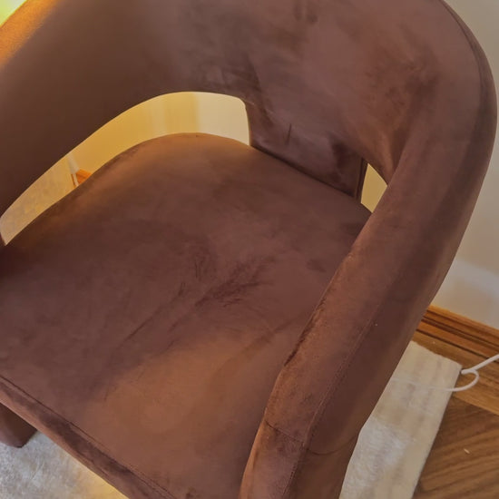 
A close-up of a modern, rust-colored armchair with a unique, open-back design and a plush, velvety texture. The camera pans out to reveal a matching chair and a small, round side table. The side table is decorated with a lit candle and a wavy-stemmed lamp with a pleated, yellow-toned shade, creating a warm and cozy atmosphere. The furniture is placed on a soft, white rug, emphasizing the room's elegant and contemporary aesthetic.