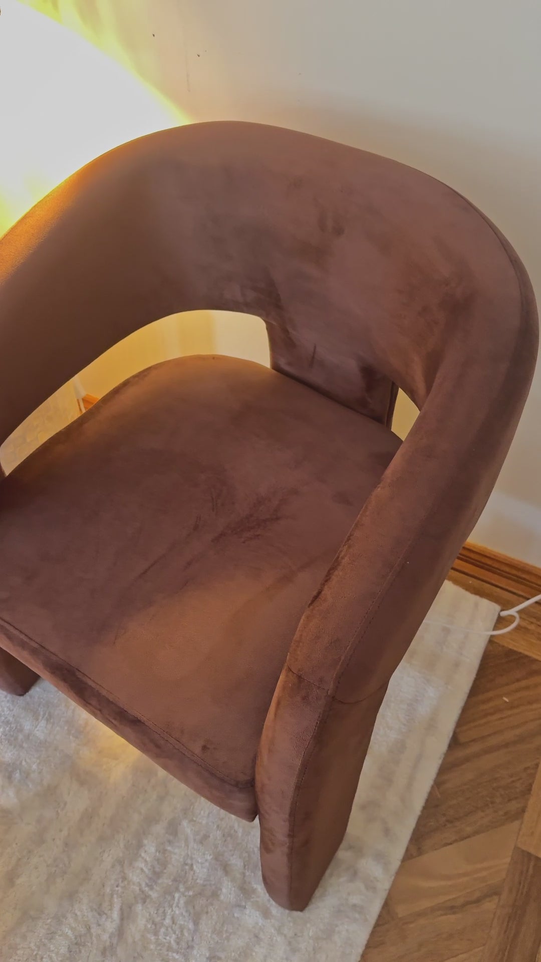 
A close-up of a modern, rust-colored armchair with a unique, open-back design and a plush, velvety texture. The camera pans out to reveal a matching chair and a small, round side table. The side table is decorated with a lit candle and a wavy-stemmed lamp with a pleated, yellow-toned shade, creating a warm and cozy atmosphere. The furniture is placed on a soft, white rug, emphasizing the room's elegant and contemporary aesthetic.