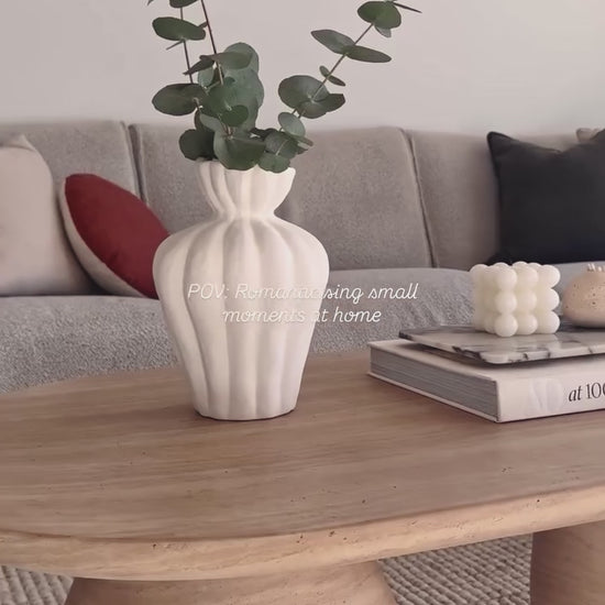 
A video showing a beautifully styled coffee table with a unique oval shape and two cylindrical legs. The camera pans slowly to reveal a white fluted vase holding eucalyptus branches and a stack of books topped with a marble tray and small, decorative candles. The table sits on a woven rug in a living room with a gray sofa and decorative pillows, showcasing a cozy, neutral aesthetic.