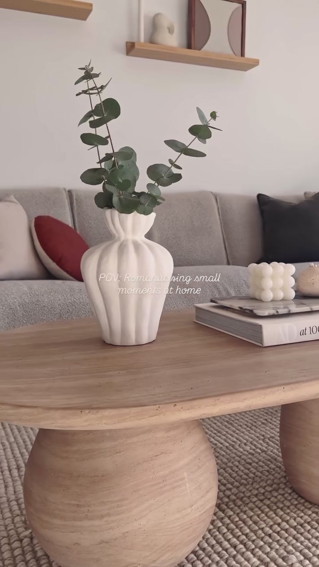 
A video showing a beautifully styled coffee table with a unique oval shape and two cylindrical legs. The camera pans slowly to reveal a white fluted vase holding eucalyptus branches and a stack of books topped with a marble tray and small, decorative candles. The table sits on a woven rug in a living room with a gray sofa and decorative pillows, showcasing a cozy, neutral aesthetic.