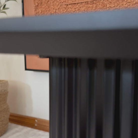 
A slow, cinematic close-up of a modern, black coffee table. The camera focuses on the table's unique base, highlighting the ridged, half-circle design of its legs. The textured surface of the legs contrasts with the smooth, rectangular tabletop. The shot then pans up to show a stack of books and a black candle holder on the table. The surrounding area includes a textured orange wall and a fluffy white rug, emphasizing the table's contemporary aesthetic.