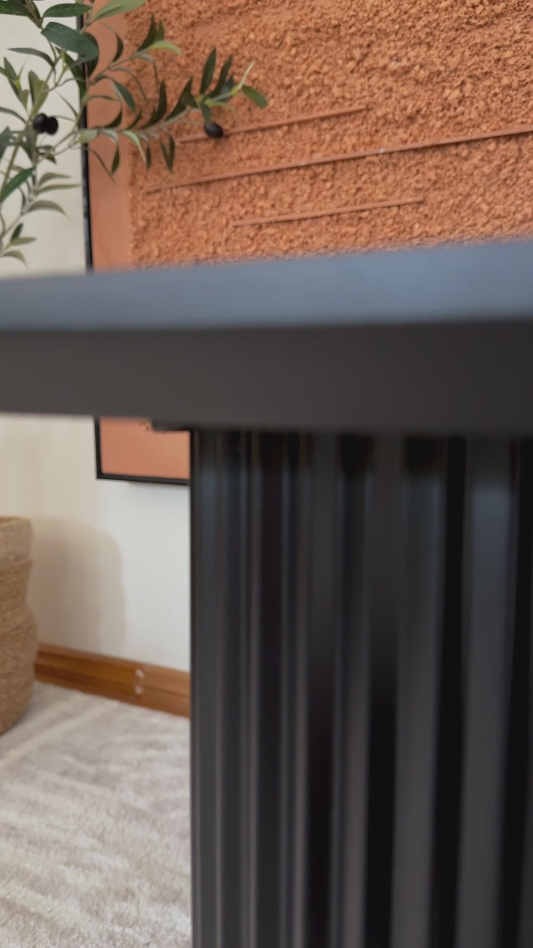 
A slow, cinematic close-up of a modern, black coffee table. The camera focuses on the table's unique base, highlighting the ridged, half-circle design of its legs. The textured surface of the legs contrasts with the smooth, rectangular tabletop. The shot then pans up to show a stack of books and a black candle holder on the table. The surrounding area includes a textured orange wall and a fluffy white rug, emphasizing the table's contemporary aesthetic.