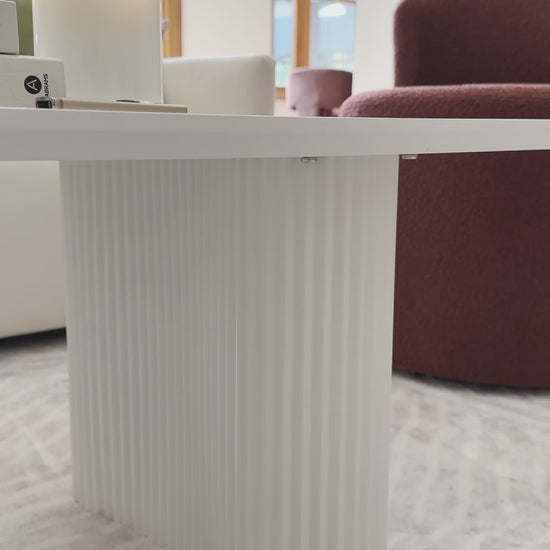 
A close-up of a modern, white coffee table with a unique rectangular design and softly rounded edges. The camera pans slowly along the table's base, which is made up of two distinct, semicircular ridged legs. The table is styled with a small houseplant, a stack of books, and a lit candle. The shot then widens to show the table on a textured rug, next to a neutral-toned sofa and a rust-colored armchair, highlighting the contemporary and cozy aesthetic of the living space.