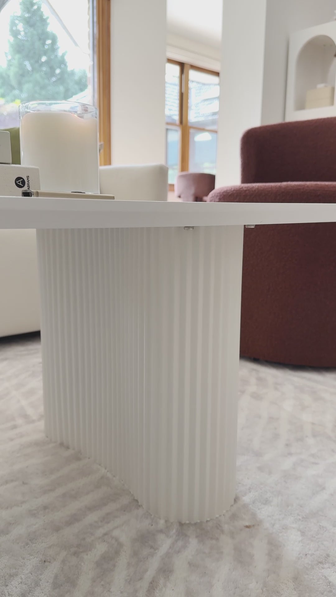 
A close-up of a modern, white coffee table with a unique rectangular design and softly rounded edges. The camera pans slowly along the table's base, which is made up of two distinct, semicircular ridged legs. The table is styled with a small houseplant, a stack of books, and a lit candle. The shot then widens to show the table on a textured rug, next to a neutral-toned sofa and a rust-colored armchair, highlighting the contemporary and cozy aesthetic of the living space.