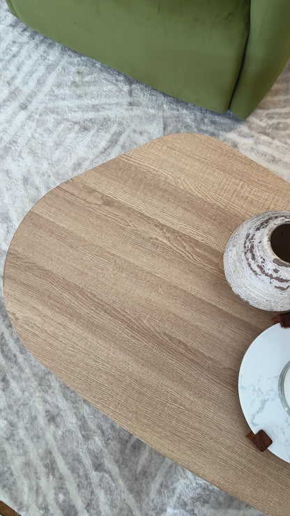 
A close-up of a modern, light oak coffee table with a freeform, organic shape. The camera slowly pans over the table's surface, showing its natural wood grain. The table is styled with two rustic vases and a lit candle on a marble tray. The shot also shows a plush, gray rug and the corner of a green velvet sofa, highlighting the cozy, inviting aesthetic of the living space.