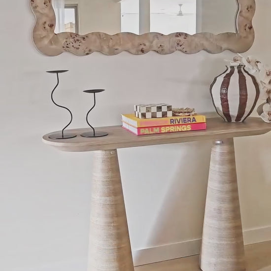 
A close-up shot of a modern console table with two conical, fluted legs and a rounded tabletop. The table is styled with a set of decorative books, a black candlestick holder, and a unique striped vase with white orchid flowers. The video then pulls back to reveal the full setting, including a large, organically shaped wooden mirror hanging above the console table.