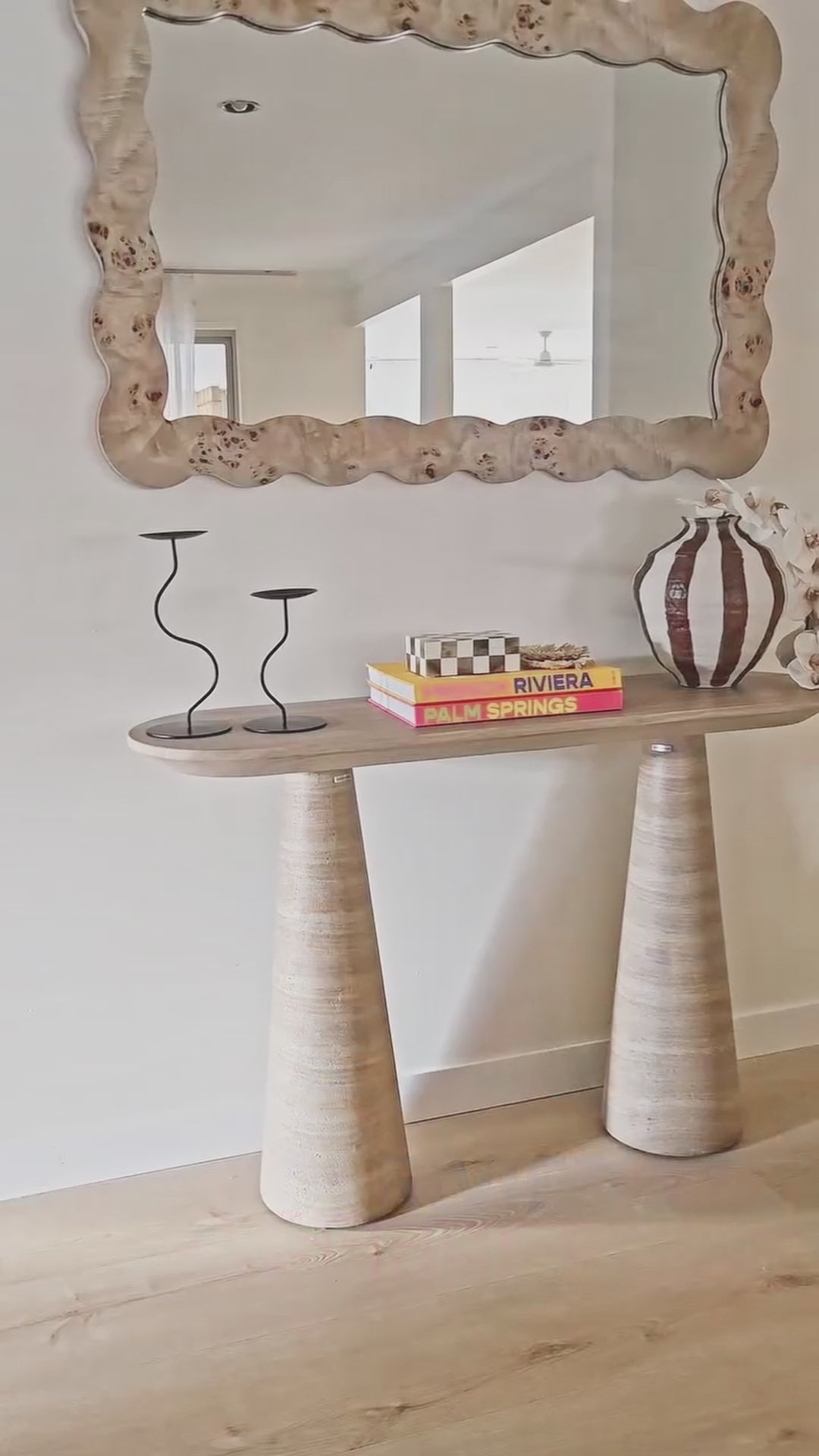 
A close-up shot of a modern console table with two conical, fluted legs and a rounded tabletop. The table is styled with a set of decorative books, a black candlestick holder, and a unique striped vase with white orchid flowers. The video then pulls back to reveal the full setting, including a large, organically shaped wooden mirror hanging above the console table.
