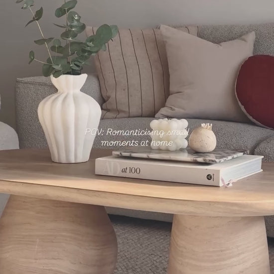 
A slow, cinematic close-up of a modern living room coffee table, showcasing its unique oval shape and a chunky, cylindrical leg. The table is styled with a white fluted vase holding green branches, a stack of books with a small marble tray, and two decorative candles. The camera slowly pans out to reveal the full living space, which includes a gray sofa with various pillows and two white, textured armchairs. The words "POV: Romanticising small moments at home" are overlaid on the video.
