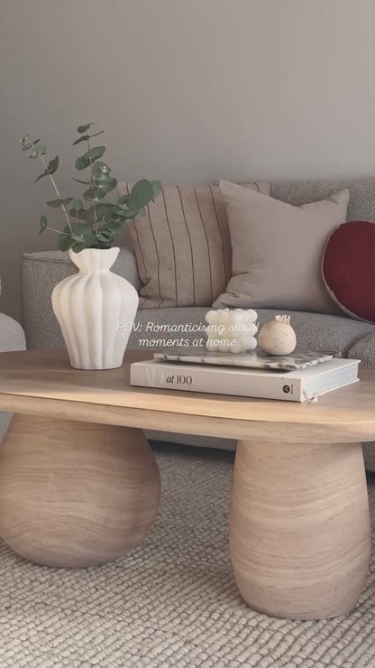 
A slow, cinematic close-up of a modern living room coffee table, showcasing its unique oval shape and a chunky, cylindrical leg. The table is styled with a white fluted vase holding green branches, a stack of books with a small marble tray, and two decorative candles. The camera slowly pans out to reveal the full living space, which includes a gray sofa with various pillows and two white, textured armchairs. The words "POV: Romanticising small moments at home" are overlaid on the video.
