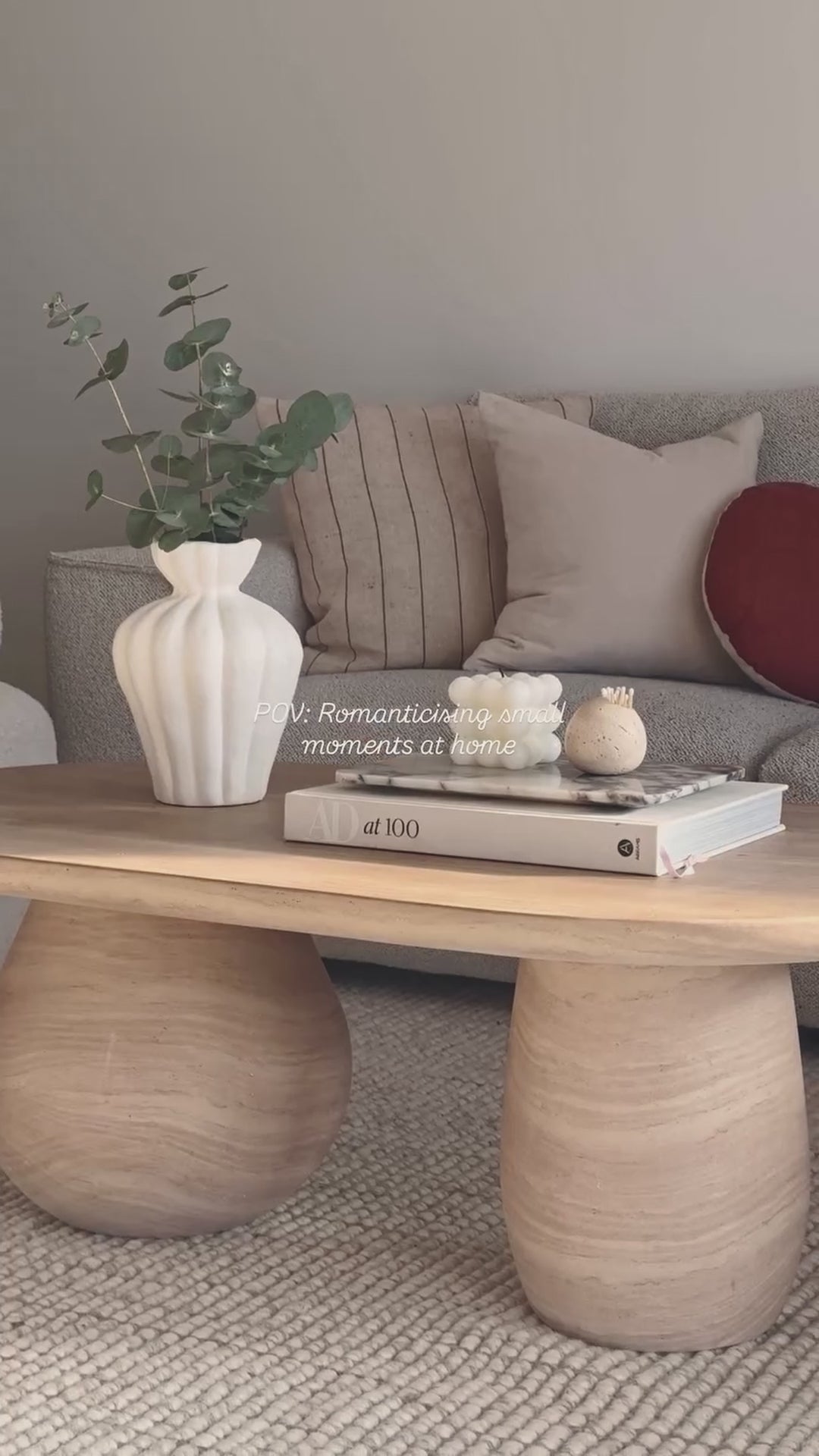 
A slow, cinematic close-up of a modern living room coffee table, showcasing its unique oval shape and a chunky, cylindrical leg. The table is styled with a white fluted vase holding green branches, a stack of books with a small marble tray, and two decorative candles. The camera slowly pans out to reveal the full living space, which includes a gray sofa with various pillows and two white, textured armchairs. The words "POV: Romanticising small moments at home" are overlaid on the video.