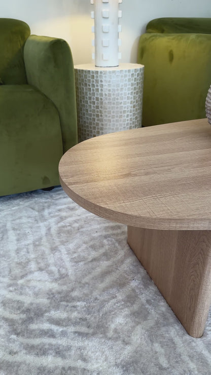 
A slow, cinematic close-up of a modern living room coffee table, showcasing its freeform, organic shape and light oak wood finish. The tabletop's natural grain is visible as the camera pans slowly across it. The table is styled with a rustic-looking vase, a larger textured vase, and a lit candle on a marble tray. The shot then pans up to show a vibrant green velvet sofa with pillows and a side table with a decorative lamp, creating a cozy and inviting atmosphere.