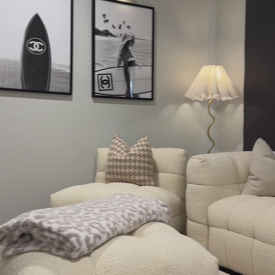 
A slow, cinematic pan over a modern living room featuring a cream-colored, textured modular sofa with decorative pillows. The camera focuses on the sofa's bouclé fabric, showing a close-up of its woven texture and tufted design. The scene includes a matching ottoman, black roller blinds, and two framed, black-and-white art prints, one of which has a Chanel surfboard. The shot highlights the furniture's texture and neutral aesthetic.