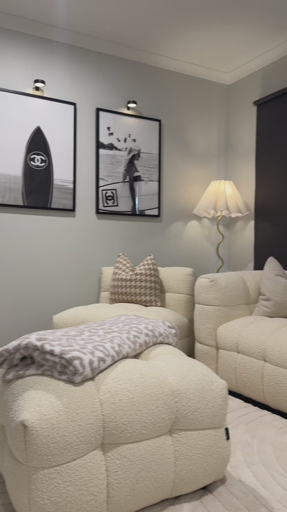 
A slow, cinematic pan over a modern living room featuring a cream-colored, textured modular sofa with decorative pillows. The camera focuses on the sofa's bouclé fabric, showing a close-up of its woven texture and tufted design. The scene includes a matching ottoman, black roller blinds, and two framed, black-and-white art prints, one of which has a Chanel surfboard. The shot highlights the furniture's texture and neutral aesthetic.
