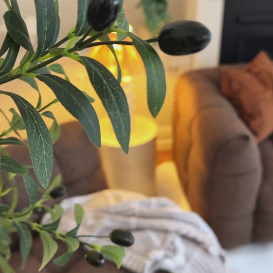 
A close-up of a leafy branch in the foreground, with a brown, textured modular sofa in the background. The camera pans out to reveal the full living room, which includes a fireplace with a mantel, built-in shelving, and a large, soft rug. The sofa is styled with an earth-toned throw blanket and two decorative pillows—one burnt orange and one with an abstract, light-colored design. A modern, wavy-stemmed lamp and a white, curved coffee table complete the cozy, contemporary aesthetic.