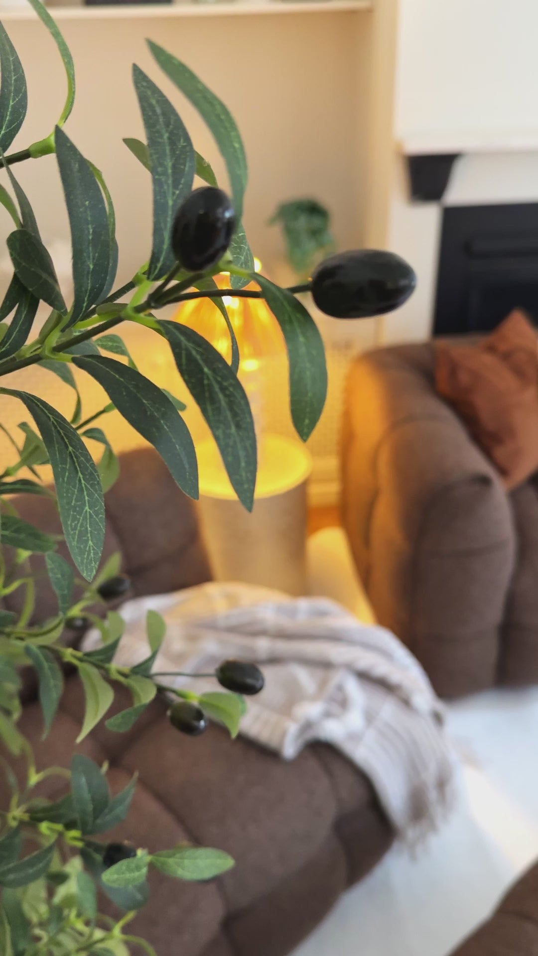 
A close-up of a leafy branch in the foreground, with a brown, textured modular sofa in the background. The camera pans out to reveal the full living room, which includes a fireplace with a mantel, built-in shelving, and a large, soft rug. The sofa is styled with an earth-toned throw blanket and two decorative pillows—one burnt orange and one with an abstract, light-colored design. A modern, wavy-stemmed lamp and a white, curved coffee table complete the cozy, contemporary aesthetic.